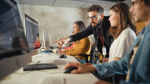 Four people work at computers in an office. A man is pointing at a monitor while talking to a woman beside him, suggesting collaboration or instruction.
