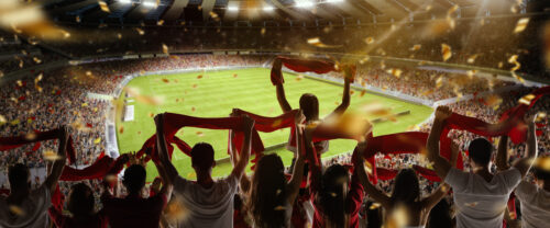 Back view of football, soccer fans cheering their team with colorful scarfs at crowded stadium at evening time. Concept of sport, support, competition. Out of focus effect