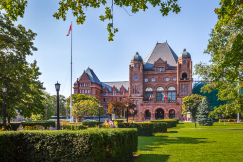 Queens Park, a large historic building with a red brick facade, arched windows, and towers, set in a green park with a flagpole and manicured hedges.